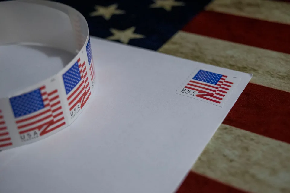 A roll of United States flag postage stamps resting on a white envelope, one stamp already applied