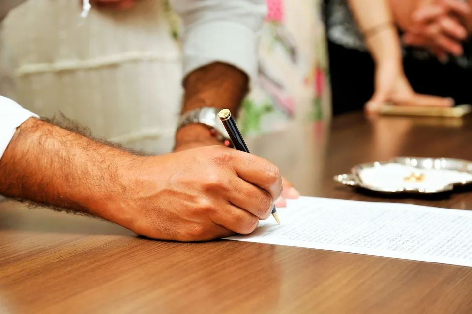 A man's hand signing a printed document with a black pen on a wooden table, a small silver tray with two rings beside it