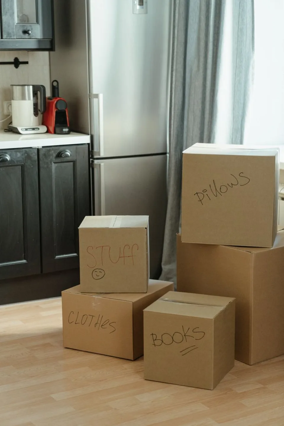 Cardboard moving boxes hand-labeled Pillows, Toys, Clothes and Books stacked on a kitchen floor