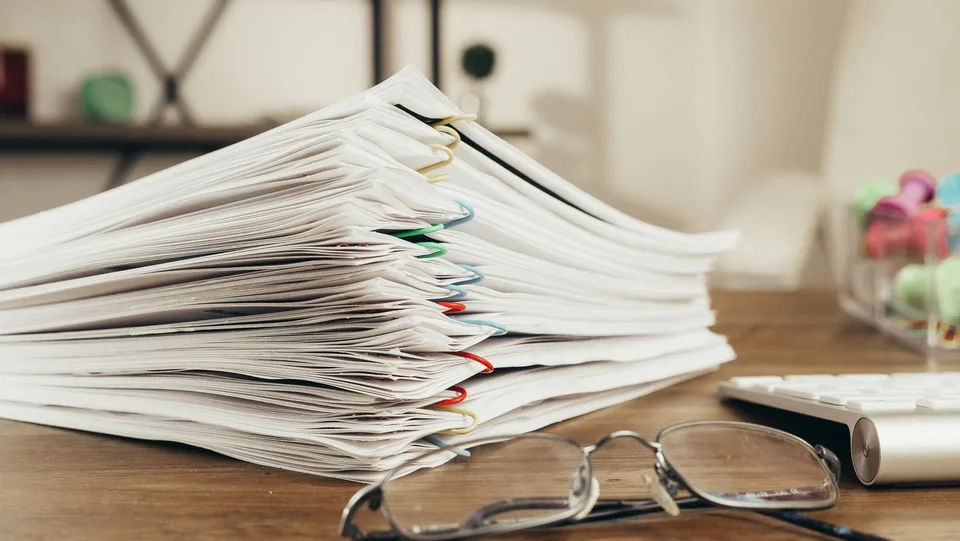A tall stack of paper documents held together with colored paper clips on a wooden desk, a pair of eyeglasses in front