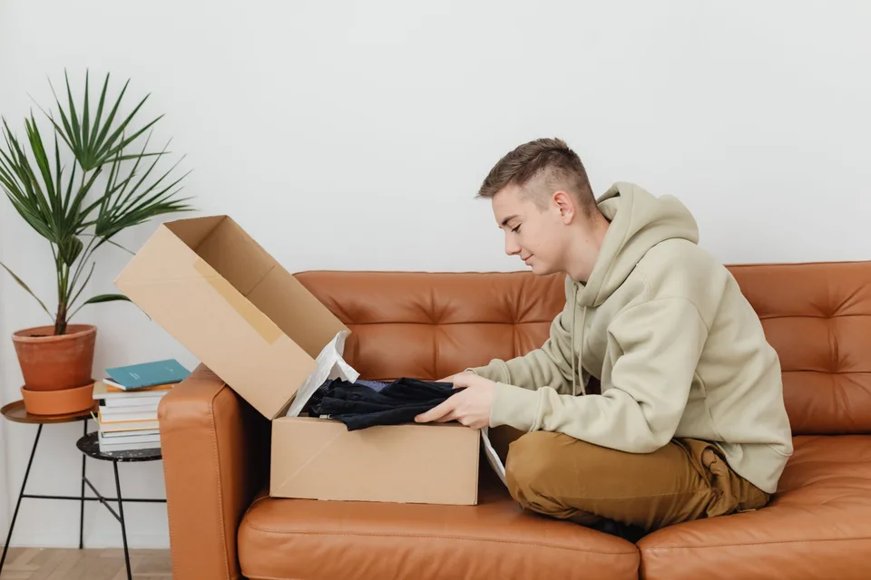A young man in a hooded sweatshirt sitting on a leather couch, lifting clothing out of an open cardboard box