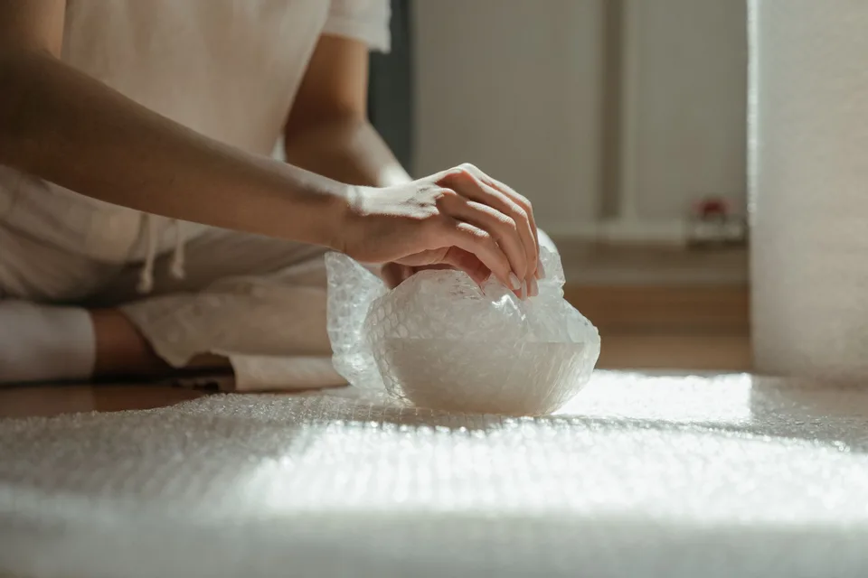 A person kneeling on the floor wrapping a white bowl in bubble wrap, a roll of bubble wrap spread out in the sunlight